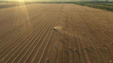 Manzaranın üstünde traktörün arka ışığı mercimeği fırına verirken, kamışı yuvarlayan ve tarımsal alanın üzerine yuvarlanmış bir balya tüküren makine, kapağı açar ve paketi dışarı atar..