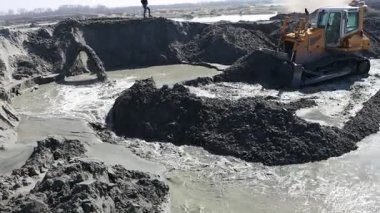 Buldozer, sürüngen kumu büyük borudaki su ve kum akışını yönlendirmek için hareket ettiriyor. Kum tortusunu boşaltmak için pompalıyor. Açılmış yüzey madenciliğinden, kazıdan ve tahsisattan su akıyor