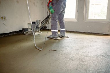 Kendi seviyesini yükselten mala parlatıcısı kullanan bir işçi. Elektrik makinesinin pedallı, plakalı tekerlekleri var ve beton levhaların üzerinde düzleştirerek daireler çiziyor..