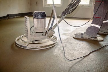 Beton levhanın yüzeyini yumuşatmak için kendi seviyesini yükselten mala parlatıcı makinesinin yakın çekimi. Benzin motoru olan bir makinenin üzerinde oturan işçinin pedli, plakalı tekerlekleri var ve onu daireler çizerek sürüyor..