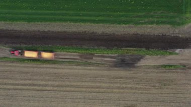 En üstte, dolly iki karavanla traktörün üzerinden uçuyor, tahıl hasadı dolu, mısırlar naklediliyor, kirli toprak yolda ilerliyor, ekilebilir tarlalar arasında yol..