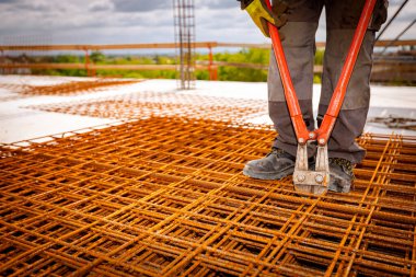 İşçi paslı dikdörtgen çelik yığınının üzerinde duruyor, beton takviyesi için inşaat alanındaki cıvata kesicilerle örgü örüyor..