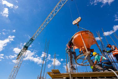 İnşaat işçileri konik metal kovaları boşaltıyorlar, taşınıyor ve beton karışımını yüksek tutuyorlar, binanın sütunları için dikey kalıba beton döküyorlar, koni tankını taşırken vinç kullanıyorlar..