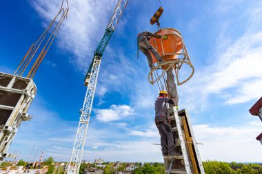 İnşaat işçileri konik metal kovaları boşaltıyorlar, taşınıyor ve beton karışımını yüksek tutuyorlar, binanın sütunları için dikey kalıba beton döküyorlar, koni tankını taşırken vinç kullanıyorlar..