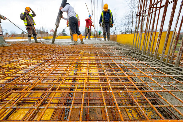 Construction worker in rubber boots is directing pump tube on right direction, pouring layer of concrete, leveling cement over square reinforcement. They using power tool, compactor, for compacting 