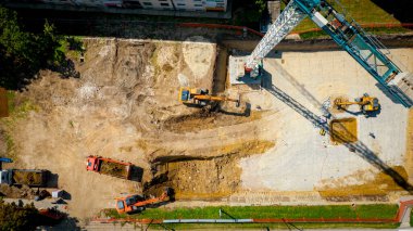 İki kazıcının kazı yaptığı, konut inşaatı için temel kazdığı ve birkaç çöp kamyonunu inşaat alanında zemin ile doldurduğu görüşünün üzerinde. Dengeli kule vinci, denge ağırlıkları