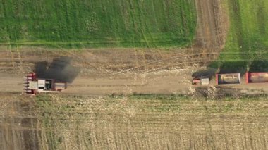 Üstten bakıldığında, doli hareketi, kırsal yoldaki tarımsal alanların arasından geçen, tozlu zemin yolunda ilerleyen hasat makinesini takip ediyor. İki tane boş römorklu traktör takip ediyor. 