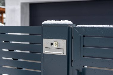 A modern videophone with a letterbox and a wireless card reader, mounted in panel fence in anthracite color, visible wicket, it rains in winter.