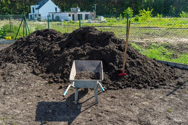 A heap of pure black earth lying in the yard next to the fence, visible shovel and empty wheelbarrow.