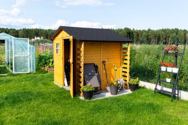 A wooden garden shed standing on a concrete foundation in a garden, flowers and tools visible. clipart