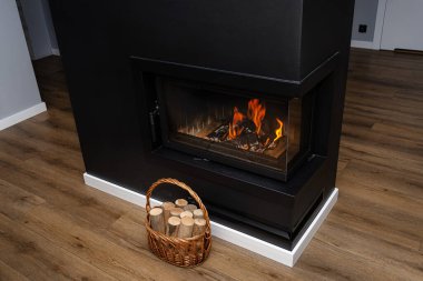 Huş ağacının parçaları hasır sepetin içinde duruyor. Kapalı bir şöminesi ve köşeli camları olan modern bir şöminenin yanında..