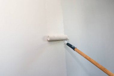 Ressam, evdeki uzatılmış bir çubukta boya silindiri kullanarak bej renkli bir oda boyuyor..