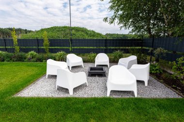 Yedi beyaz plastik sandalye kaba çakılların üzerindeki metal bir ateş çukurunun etrafına dizilmiştir..