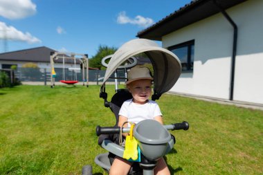 On beş aylık bir bebek evin yanındaki çimenlikte üç tekerlekli bisiklete biner..