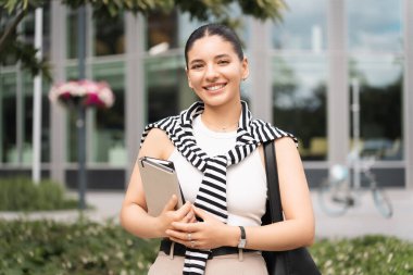 Beyaz bir atlet ve çizgili süveter giymiş gülümseyen genç bir kadın bir çanta ile şehirde yürüyor. Öğrenci ve iş kadını rollerini dengeleyen modern profesyonelin vücut bulmuş halidir..