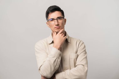 Derin düşüncelere. Gözlüklü ve bej gömlekli bir adam çenesini tutuyor, düşünceli bir şekilde bakıyor. Zeka ve modern stilin mükemmel bir karışımı..