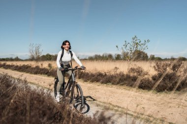 Hafif giysiler içinde gülümseyen bir kadın bisiklet sürüyor, sağlık, enerji saçıyor ve açık havaya karşı saf bir sevgi besliyor..