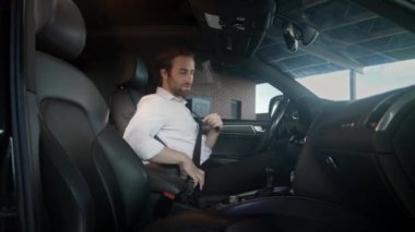 Beyaz gömlekli bir adam sürücü koltuğuna kayıyor. Kapıyı kapattı ve emniyet kemerine uzandı. Her şeyin yolunda olduğundan emin olunca motoru çalıştırır ve yola çıkmaya hazırlanır..