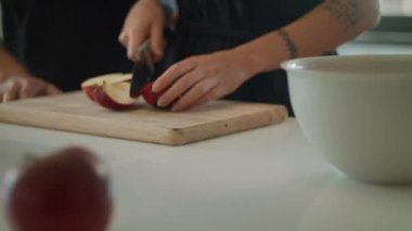Dövmeli bir kadın kırmızı elmayı çeyreklik şeklinde keser. Kesme tahtası ve bıçak kullanıyor. Parçaları beyaz bir kaseye koyar. Diğer elmalar masanın üstünde. Yanında bir erkek eli görüyoruz..
