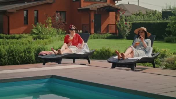 女朋友们正坐在阳光下悠闲自在地聊天 他们前面有一个游泳池 姑娘们戴着太阳镜 他们正在喝鸡尾酒 看起来很放松 — 图库视频影像