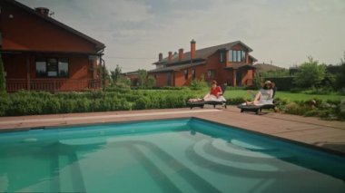 İki zarif giyimli genç kadın, birbirlerine kızarak konuşmayı bitiriyor. Güneş pansiyonlarında oturuyorlar, önlerinde bir yüzme havuzu var. Arka planda iki lüks ev var..