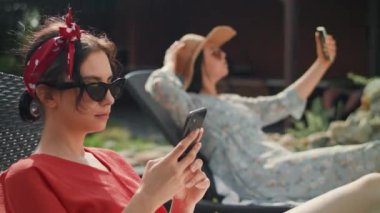 Kamera kırmızı bluzlu, güneş gözlüklü bir kadına odaklanmış. Bir güneş şezlonguna oturuyor ve akıllı telefonunu kullanıyor. Mavi elbiseli genç bir kadın daha var. Telefonuyla selfie çekiyor..