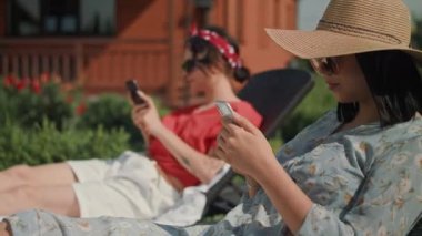 Çiçekli elbiseli genç bir kadın güneş panelinde otururken telefonuna bakar. İkinci güneş pansiyonunda bir kadın daha var. Ayrıca telefonunu da karıştırıyor. Kolunda bir dövme var..