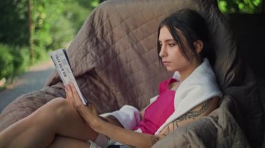 Dövmeli kollu genç bir kadın sallanan koltukta otururken kitap okuyor. Esniyor ve uyumak istiyor. Dışarıda kırmızı bir bluz ve omuzlarında beyaz bir havluyla oturuyor..