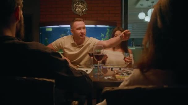 两个女人和两个男人坐在一张桌子旁玩棋盘游戏 他们似乎对此非常感兴趣 桌上有几杯酒 后面是一个水族馆 — 图库视频影像