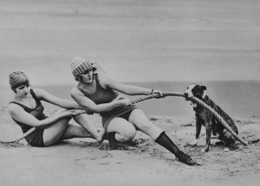 Plajda mayo giyen genç kadınlar köpekten ip çekiyorlar.