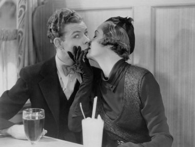 Genç bir kadın restoranda yemek masasında oturan adamı öpüyor. 