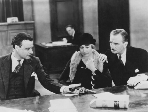 Policeman showing pistol to worried mature woman sitting with husband at desk in police station 