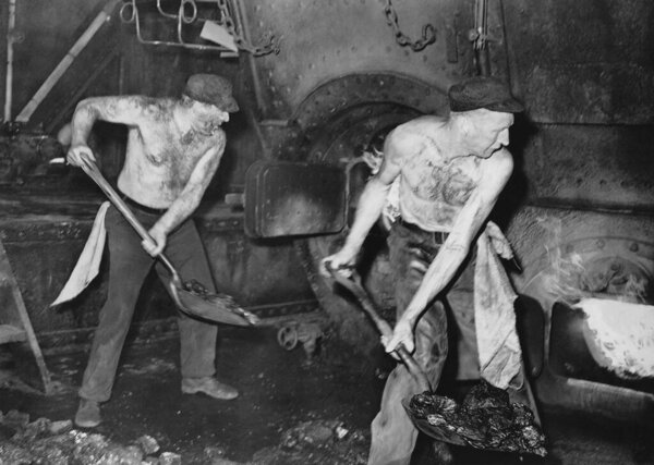 Dirty shirtless men throwing coal in furnace inside boiler room of ship 