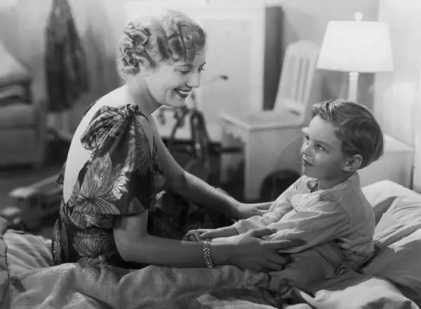 笑顔の母親は自宅でベッドに座っているかわいい息子の腕に触れる ストック画像