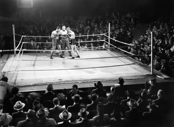 アリーナで観客に囲まれた照らされたボクシングリングのボクサーと審判 ストックフォト