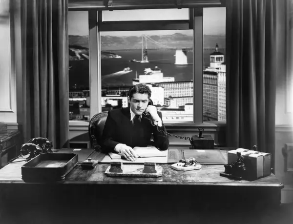 商人坐在办公室的桌子边 一边用固定电话交谈 图库照片