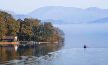 LAKE DISTRICT, UK - October 12, 2010. Boat house on shore of Coniston Water with couple paddling a double canoe. Misty morning in autumn. clipart