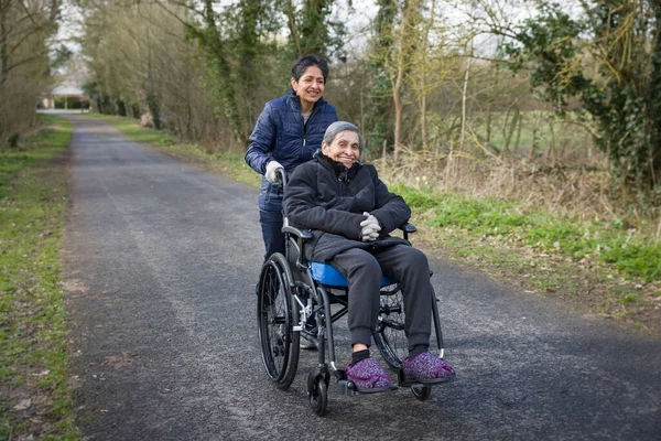 アジアのインド人女性は 冬のイギリスの屋外の車椅子で彼女の年配の母親を押します キャリアを描写し コミュニティでケアすることもできます — ストック写真