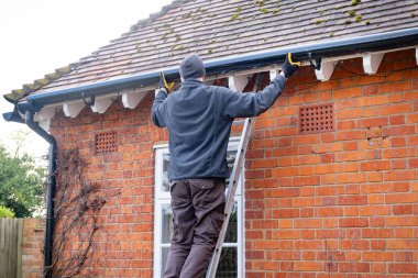 Man on a ladder fixing fitting a rain gutter on the eaves of a UK house clipart