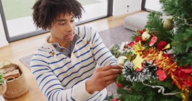 Ren geyiği boynuzlu mutlu bir adamın evde Noel ağacı süslediği video. Noel, kutlama, gelenek, mutluluk ve kapsayıcılık kavramı.