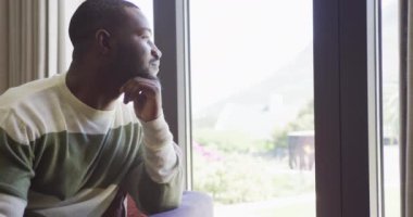 Düşünceli Afro-Amerikan bir adamın evde oturup pencereden dışarı baktığı bir video. Ev hayatı ve kapsayıcılık kavramı.