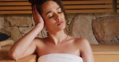Havlu giymiş, gözleri kapalı, saunada, sağlık merkezinin saunasında dinlenen melez bir kadının videosu. Tatil, rahatlama, sağlık ve kapsayıcılık kavramı.