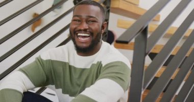 Mutlu Afro-Amerikan bir adamın video portresi kameraya gülüyor, evdeki merdivenlerde oturuyor. Ev hayatı, sağlık, mutluluk ve kapsayıcılık kavramı.