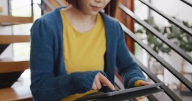 Mutlu Asyalı bir kadının gülümsediği, merdivenlerde tablet kullanarak oturduğu ve evine baktığı bir video. Ev hayatı, iletişim, sağlık, mutluluk ve kapsayıcılık kavramı.