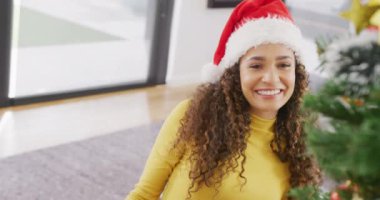 Noel Baba şapkalı mutlu kadın videosu. Gülümsüyor ve evde Noel ağacı süslüyor. Noel, kutlama, iletişim, mutluluk ve kapsayıcılık kavramı.