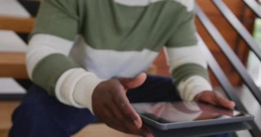 Mutlu Afro-Amerikan bir adamın güldüğü, elinde tabletle merdivenlerde oturduğu bir video. Ev hayatı, iletişim, sağlık, mutluluk ve kapsayıcılık kavramı.
