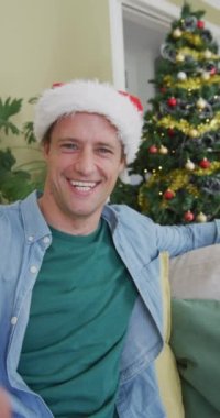 Kafkasyalı bir adamın video görüntüsü, Noel Baba şapkası takması, arkasından Noel ağacını göstermesi. Noel konsepti.