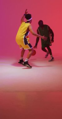 Pembe arka planda topla oynayan iki farklı erkek basketbolcunun dikey videosu. Spor ve rekabet konsepti.