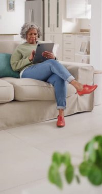 Afro-Amerikan kadının tablet kullanarak koltukta oturduğu dikey video. Emeklilik ve ev kavramında zaman geçirme.