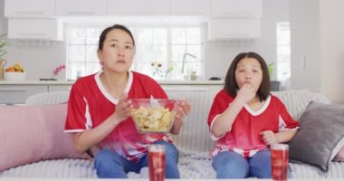 Mutlu Asyalı anne ve kızının cips yiyip TV 'de maç izlediği bir video. Aile, annelik, ilişkiler ve birlikte kaliteli zaman geçirme konsepti dijital olarak oluşturulmuş video.